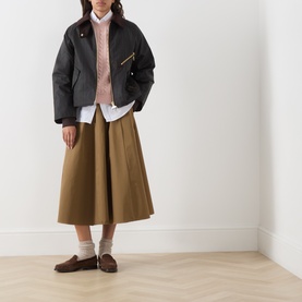 Model wearing Barbour waxed cotton jacket in Classic Shadow, styled with tan midi skirt, pink jumper, white shirt and brown loafers