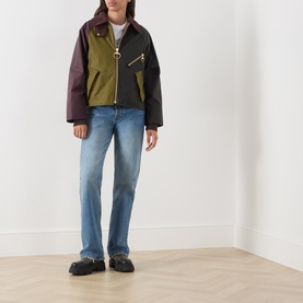 Model wearing Barbour jacket in Golden Olive/Fern colourblock waxed cotton with gold-tone zip, styled with blue straight-leg jeans and black shoes.