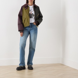Model wearing Barbour jacket in Golden Olive/Fern colourblock waxed cotton, styled with white graphic tee and blue straight-leg jeans.