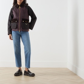 Model wearing Barbour wax cotton jacket in Rustic/Bordeaux/Muted with gold-tone hardware, styled with blue straight-leg jeans and black loafers.