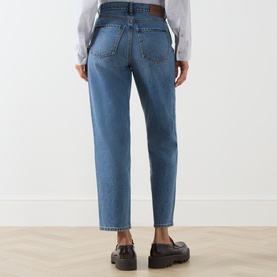 Model wearing Barbour mid blue cotton jeans, rear view, styled with a white shirt and black loafers.