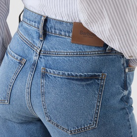 Model wearing Barbour mid blue cotton jeans, rear view showing back pockets and brown leather logo patch, styled with a white striped shirt.