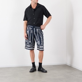 Model wearing Dolce & Gabbana navy silk shorts with white graphic print, styled with black short-sleeve shirt, black socks and loafers.