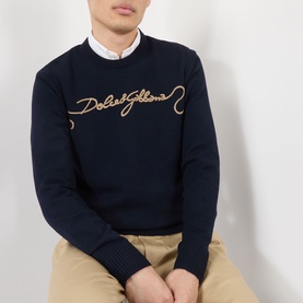 Model wearing Dolce & Gabbana dark blue wool sweatshirt with gold embroidered logo script, styled with white shirt and beige trousers