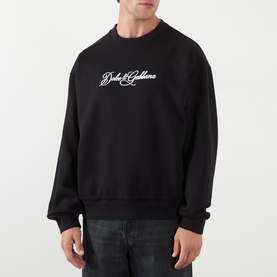Model wearing Dolce & Gabbana Logo Graphic Sweatshirt in black with contrast script logo, crew neck, ribbed trims, styled with dark jeans.
