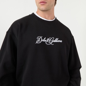 Model wearing Dolce & Gabbana Logo Graphic Sweatshirt in black, crew neck with embroidered script logo across chest, ribbed trim detail visible.