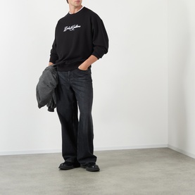 Model wearing Dolce & Gabbana black Logo Graphic Sweatshirt with dark bootcut jeans and black loafers, carrying a grey jacket