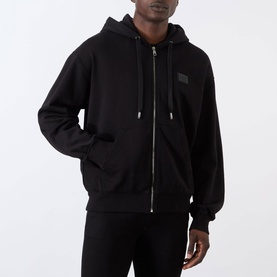 Model wearing Dolce & Gabbana black Logo Plate Hoodie with zip fastening, drawstring hood, and chest logo plate, styled with black joggers.