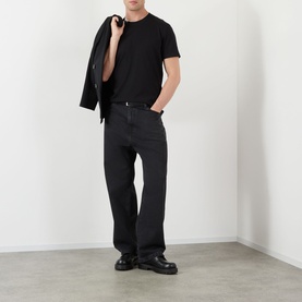 Model wearing Dolce & Gabbana Black Logo Tab T-Shirt with black trousers and chunky leather shoes, jacket draped over shoulder.