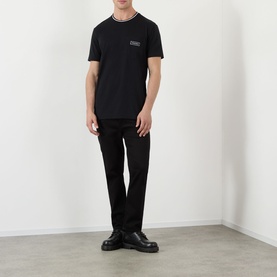 Model wearing Dolce & Gabbana black cotton T-shirt with tipped rib crew neck and logo patch, styled with black trousers and chunky leather shoes.