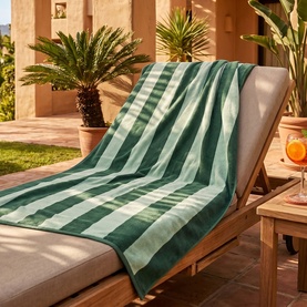 Misona Beach Towel Green Blue draped over a wooden sun lounger outdoors, featuring bold green and sage wide stripes, 100% cotton jacquard