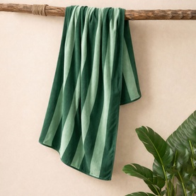 Misona Beach Towel in green blue, 100% cotton jacquard with tonal stripe pattern, draped over a wooden rail with tropical leaf backdrop.