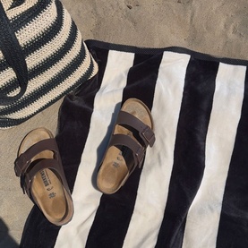 Misona Beach Towel Natural Black, 100% cotton jacquard with bold black and white stripes, styled on sand with sandals and striped bag.