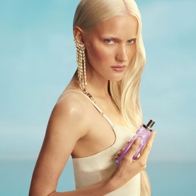 Model with Mugler Body Mist in purple bottle, wearing gold chain earrings and nude bandeau top, against blue sky background