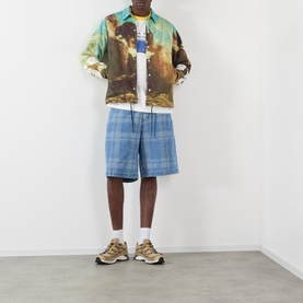 Model wearing Pleasures multi-coloured printed casual jacket, styled with blue check shorts, white tee and tan trainers.