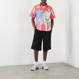 Model wearing Pleasures More Songs About Buildings and Food Check Shirt in red graphic print, styled with black shorts and white trainers.