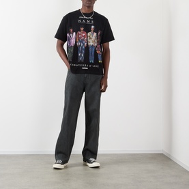 Model wearing Pleasures Creatures of Love Logo Graphic T-Shirt in black, styled with dark grey wide-leg trousers and black low-top trainers.