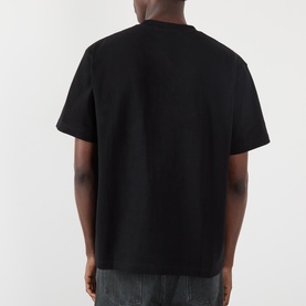 Model wearing Pleasures Creatures of Love Logo Graphic T-Shirt in black, rear view, crew neck, short sleeves, relaxed fit, styled with grey trousers.