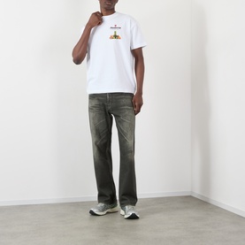 Model wearing Pleasures Delivery Back Graphic Logo T-Shirt in white, styled with dark khaki jeans and grey trainers.