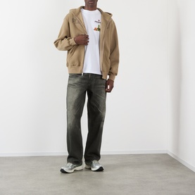 Model wearing Pleasures Delivery Back Graphic Logo T-Shirt in white, styled with a tan zip-up hoodie, dark grey jeans, and grey trainers.