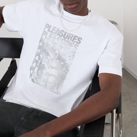 Model wearing Pleasures Immolate Logo Graphic T-Shirt in white with silver repeated logo print, styled with dark jeans and gold chain necklace.