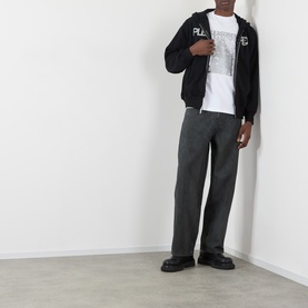 Model wearing Pleasures Immolate Logo Graphic T-Shirt in white, styled with black zip-up hoodie, grey trousers and black chunky loafers.
