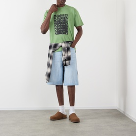 Model wearing Pleasures Immolate Logo Graphic T-Shirt in sage, styled with light blue denim shorts, plaid shirt tied at waist, and tan slippers.