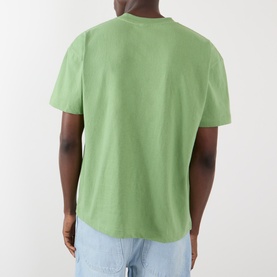 Model wearing Pleasures Immolate Logo Graphic T-Shirt in sage, rear view, short sleeves, styled with light blue jeans.