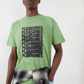 Model wearing Pleasures Immolate Logo Graphic T-Shirt in sage with dark repeat logo print, styled with checked trousers.