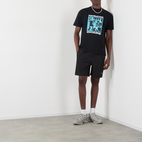 Model wearing Pleasures Navigating Chaos Logo Graphic T-Shirt in black with blue graphic print, styled with black shorts and grey trainers.