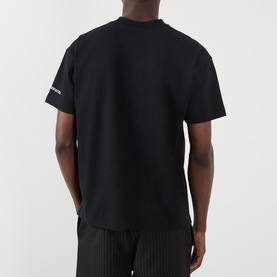 Model wearing Pleasures Navigating Chaos Logo Graphic T-Shirt in black, rear view, with striped trousers. Short sleeves, crew neck.