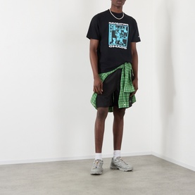 Model wearing Pleasures Navigating Chaos Logo Graphic T-Shirt in black with green plaid shirt tied at waist, black shorts and grey trainers.