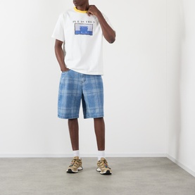 Model wearing Pleasures Speaking Logo Graphic T-Shirt in white with blue check shorts and gold-tone trainers.
