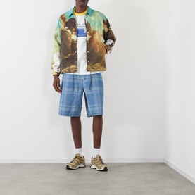Model wearing Pleasures Speaking Logo Graphic T-Shirt in white, styled with a multicolour printed jacket, blue plaid shorts, and tan trainers.