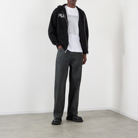 Model wearing Pleasures black cotton jeans with wide-leg fit, styled with black zip hoodie, white graphic tee and black chunky loafers