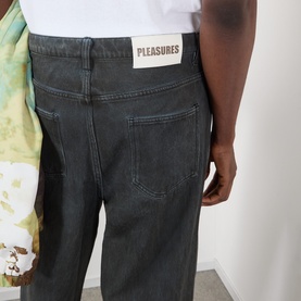 Model wearing Pleasures black cotton jeans, rear view showing branded patch label and five-pocket styling, with graphic tee visible at side.