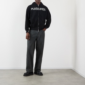 Model wearing Pleasures black wide-leg jeans in 100% cotton, styled with a black branded zip-up hoodie and chunky black shoes.