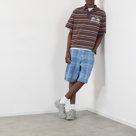Model wearing Pleasures brown striped polo shirt with embroidered chest logo, styled with blue denim shorts and grey chunky trainers.