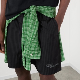Model wearing Pleasures Active Striped Shorts in black with elasticated waist and logo detail, styled with a green plaid shirt tied at waist.