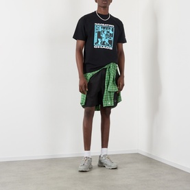 Model wearing Pleasures Active Striped Shorts in black with elasticated waist, styled with a graphic tee and tied green plaid shirt.