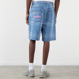 Model wearing Pleasures blue denim shorts, rear view showing pink embroidered logo on back pocket, styled with grey trainers.