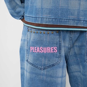 Model wearing Pleasures blue denim shorts with pink embroidered logo pocket, styled with a studded brown belt and blue check shirt.