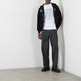 Model wearing Pleasures Zine Logo Zip Hoodie in black, styled with grey trousers and a white graphic tee.