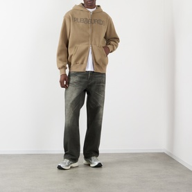 Model wearing Pleasures Zine Logo Zip Hoodie in beige, styled with dark wash wide-leg jeans and grey trainers.