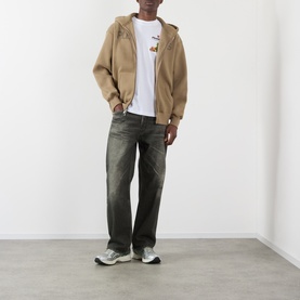 Model wearing Pleasures Zine Logo Zip Hoodie in beige with white graphic tee, dark wash jeans and grey trainers.