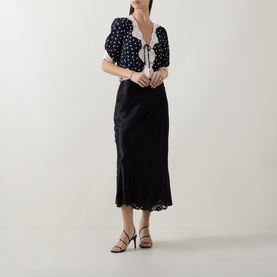 Model wearing Rixo London black viscose midi skirt with lace hem trim, styled with a black and white polka dot blouse and black heeled sandals.