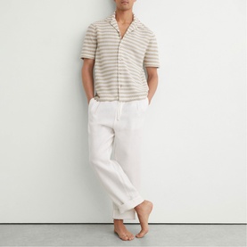 Model wearing Reiss white linen trousers with drawstring waist, styled with a beige striped short-sleeve shirt, barefoot against a white wall