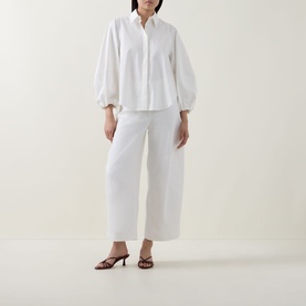 Model wearing Akris Punto cream shirt with balloon sleeves and button front, styled with white wide-leg trousers and brown heeled sandals.
