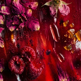 Rich red flat-lay with dried rose petals, raspberries, flower blooms, and scattered botanicals suggesting a berry-floral fragrance or beauty product