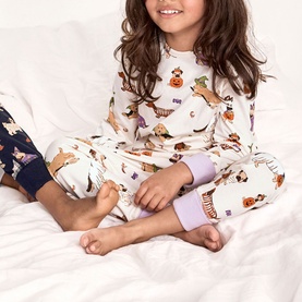 Model wearing Chelsea Peers Halloween Dogs Multi pyjama set with all-over dog costume print, lilac cuffs, on white bedding